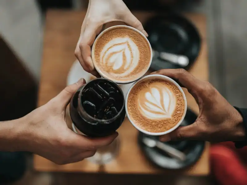 Flat White Coffee: Your Super Easy Guide to This Tasty Drink Hands holding flat white coffee cups with latte art in a cheers gesture on a wooden table.