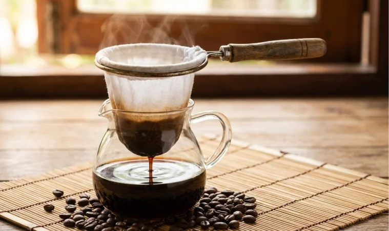 Traditional Thai coffee sock filter Tung Dtom with white cotton cloth bag and wooden handle brewing dark Oliang coffee into glass carafe on bamboo mat